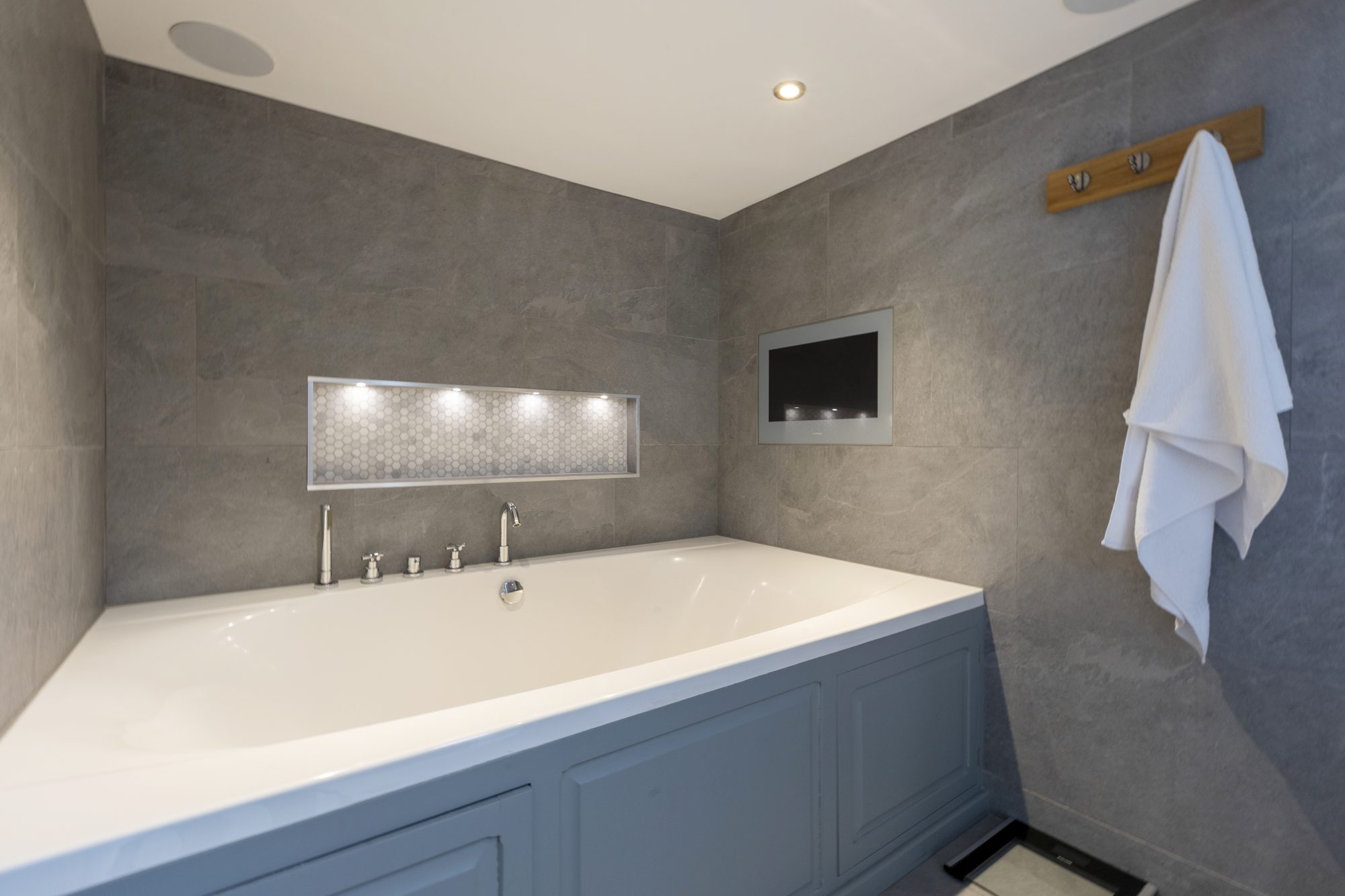 Grey-panelled bath under a lit mirror shelf
